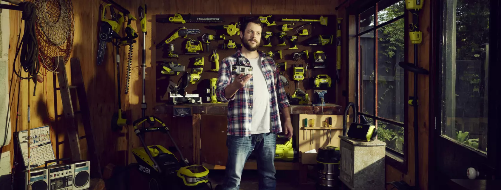 A man stands holding an 18V battery, surrounded by compatible Ryobi power and garden tools.